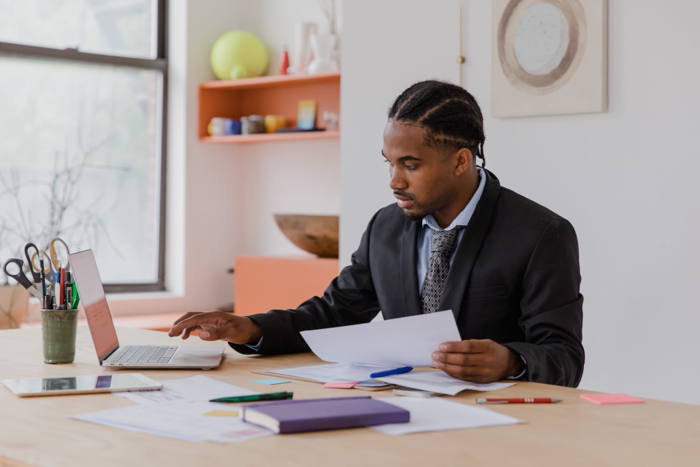 Business owner at a bright wooden desk reading through a printed quote, with a laptop, tablet and paperwork spread out in front of them
