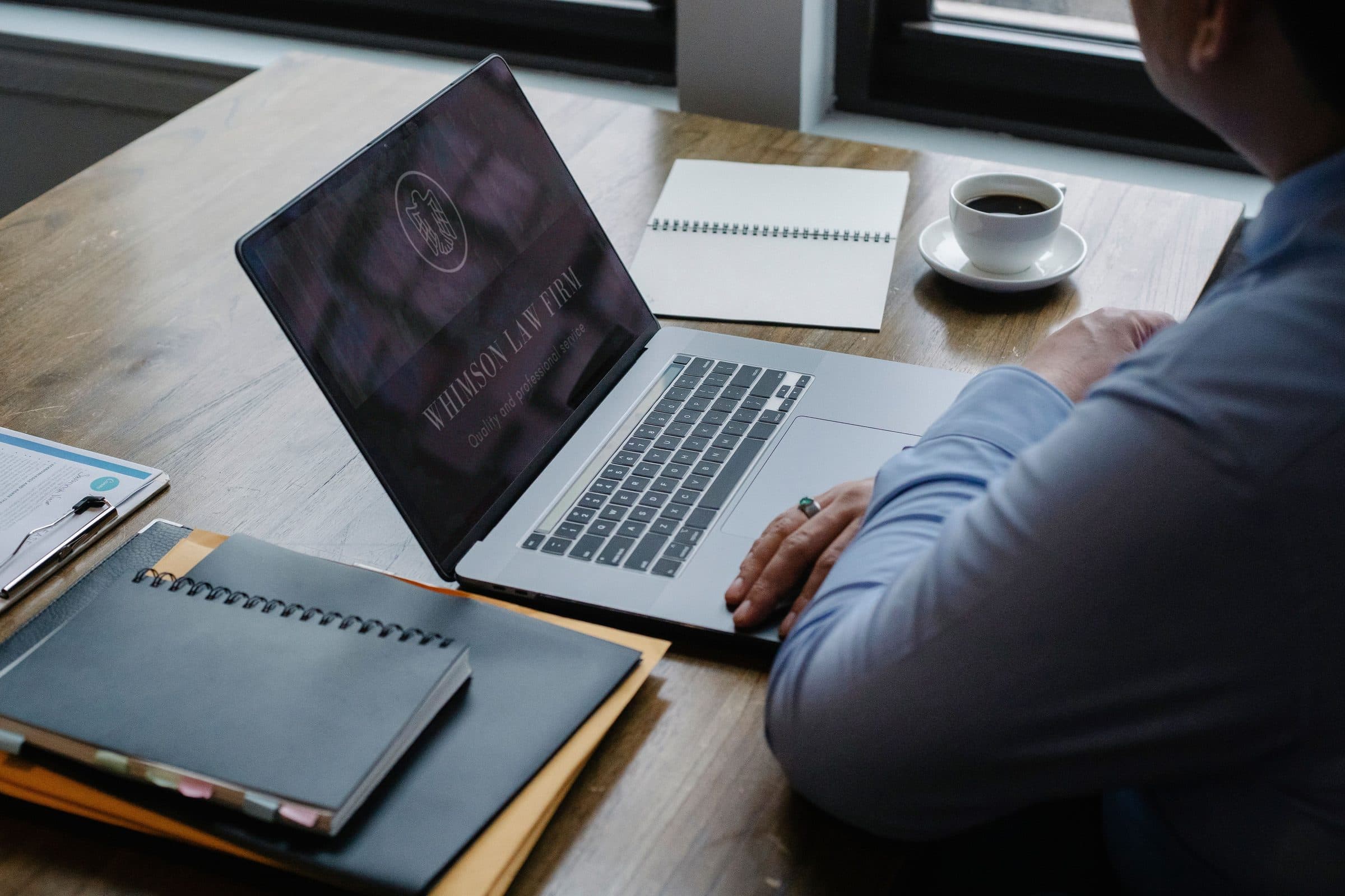 Solicitor in a blue shirt at a wooden desk looking at a law firm website on a laptop, with case files, a notebook and a cup of coffee beside it