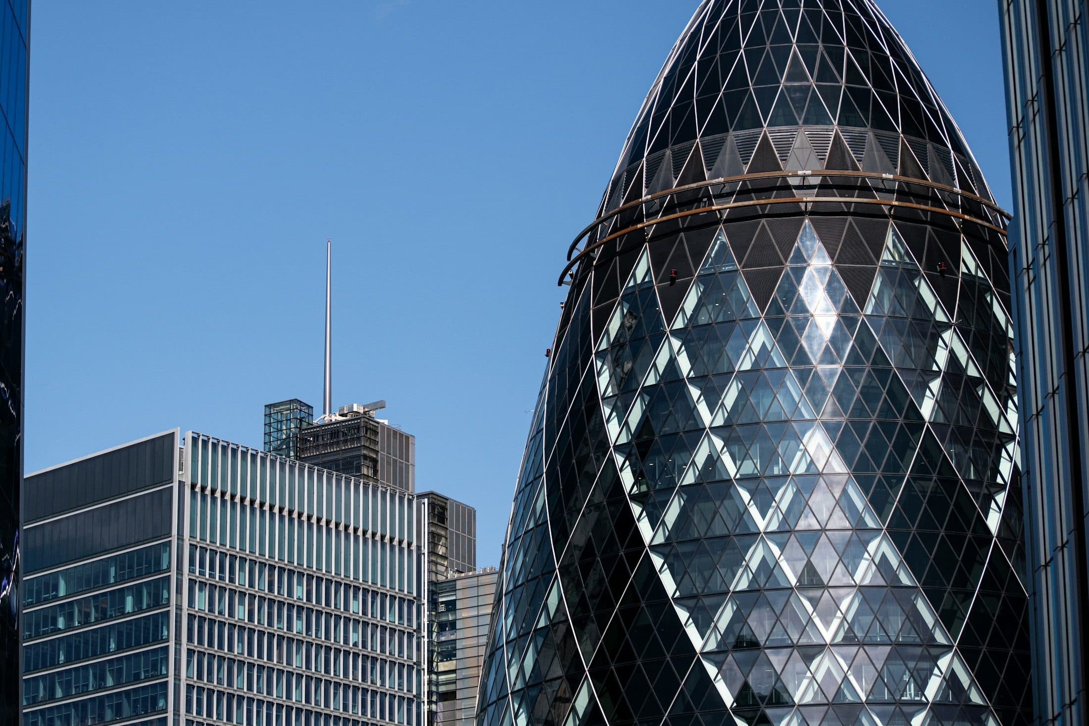 The Gherkin between two glass office towers in the City of London under a blue sky