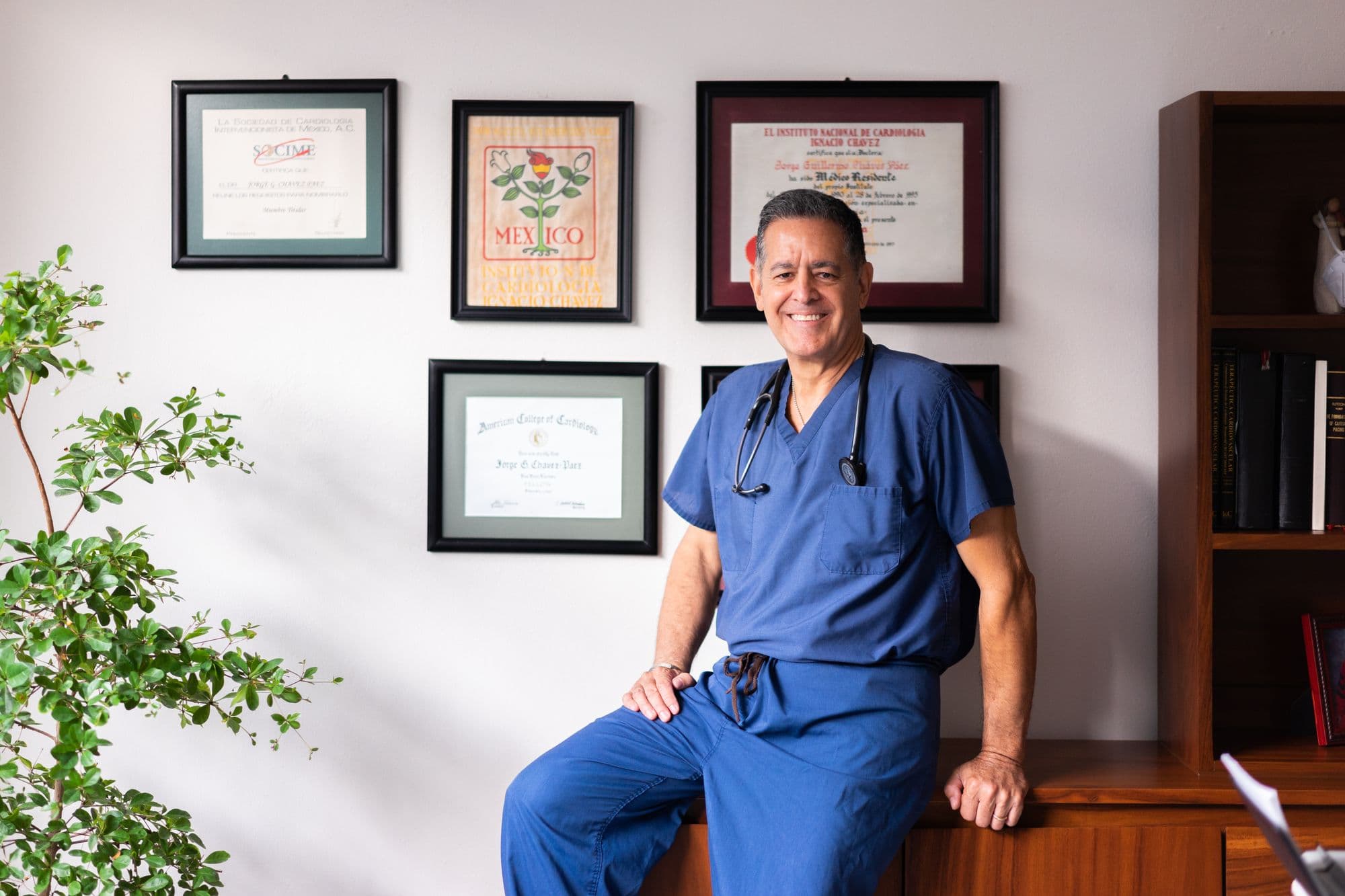 A private consultant sitting beside framed medical qualifications in a consulting room