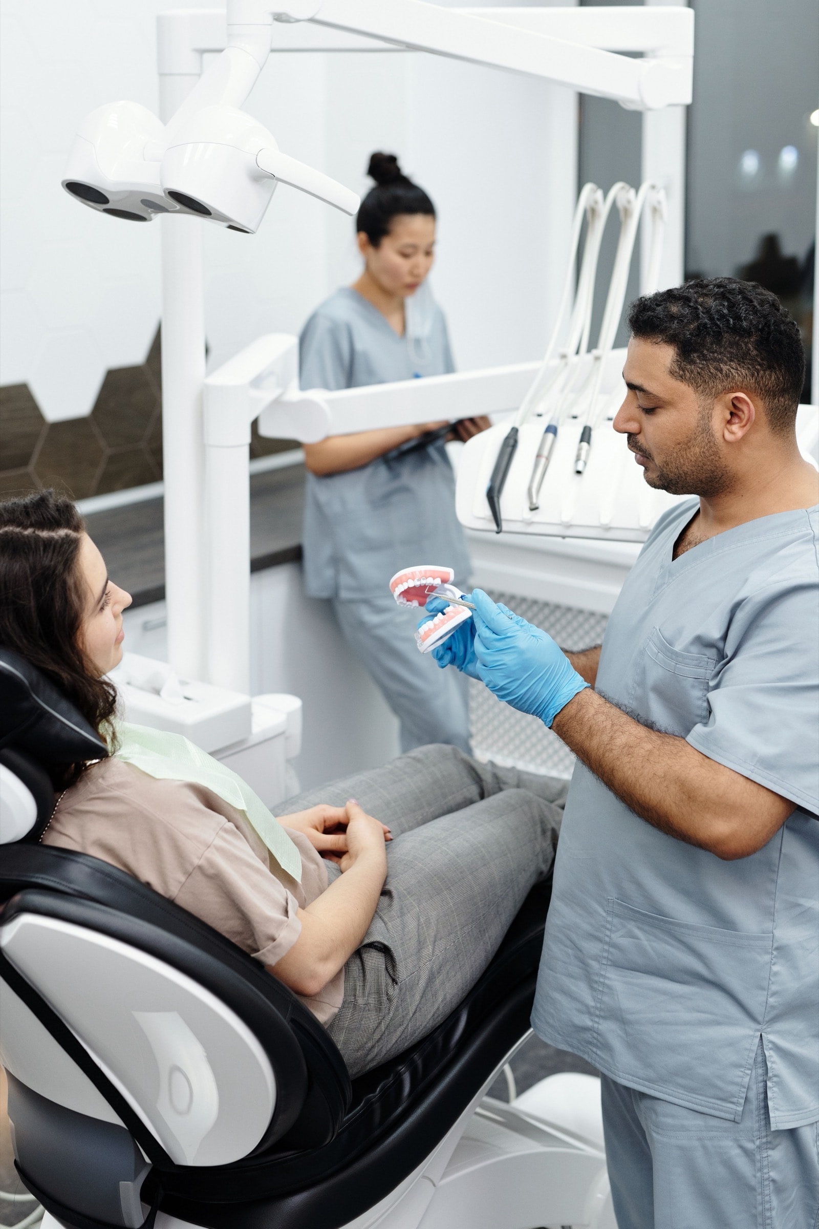 A dentist in blue scrubs showing a patient a model of the teeth, with a colleague standing behind them
