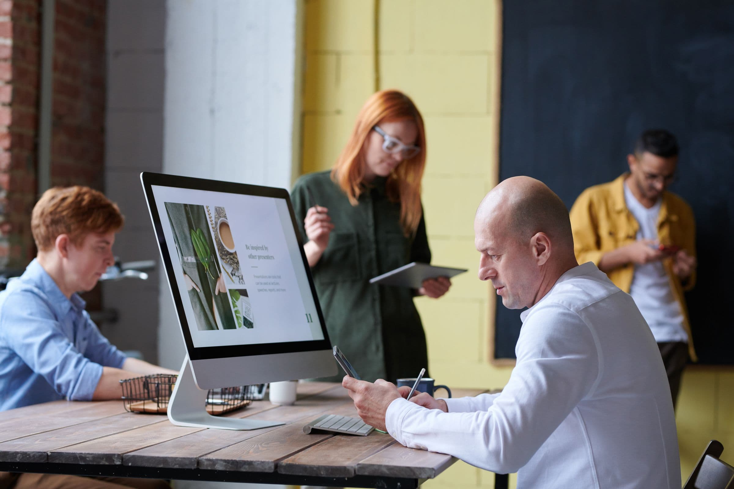 People working at a shared wooden table in a bright open-plan workspace, with a large monitor showing a presentation and a man making notes beside it