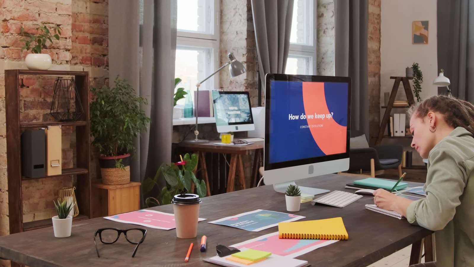 A designer sketching page ideas at her desk in a bright studio with brick walls and plants