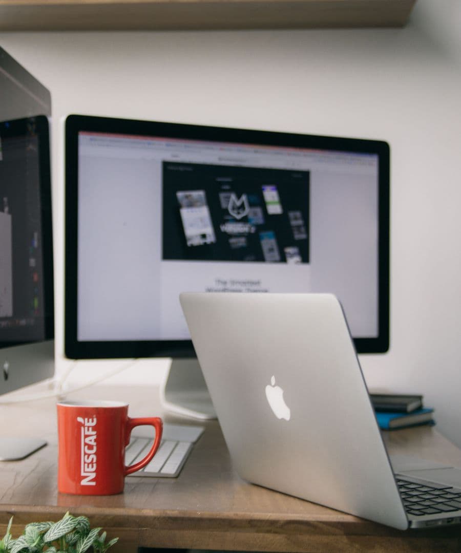 A web design studio desk with a website being built on a large monitor beside a laptop and a red mug