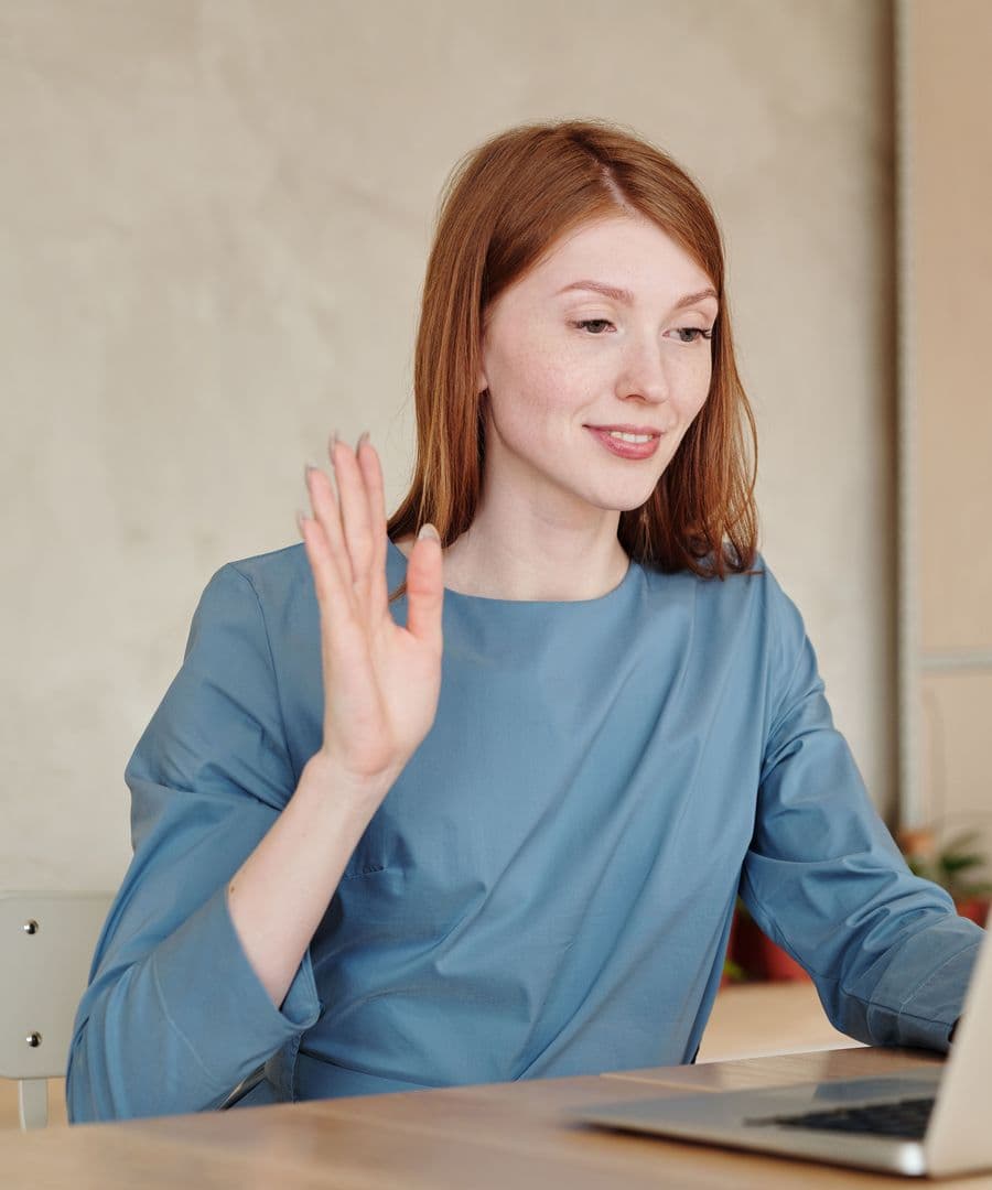 A woman smiling and waving hello at the start of a video call on her laptop