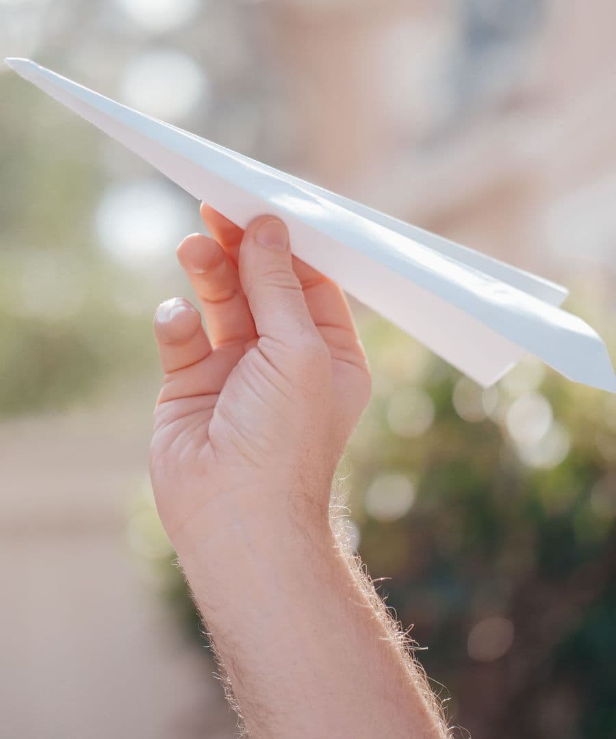 A hand holding up a white paper plane, ready to launch it, in soft outdoor light