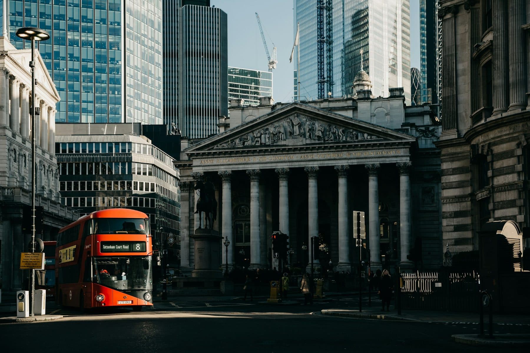 The portico of the Royal Exchange at Bank in the City of London, with a red bus in sunlight and office towers behind