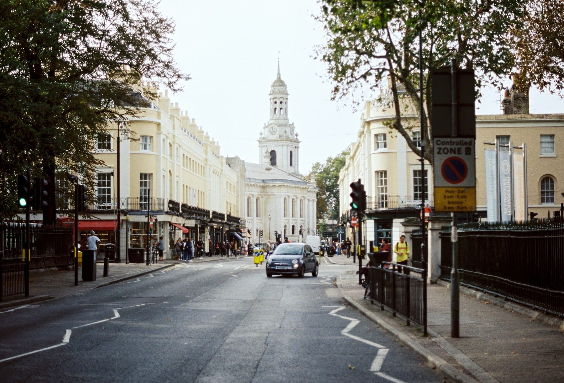 A Greenwich street of pale stucco terraces and shops with a church tower at the far end