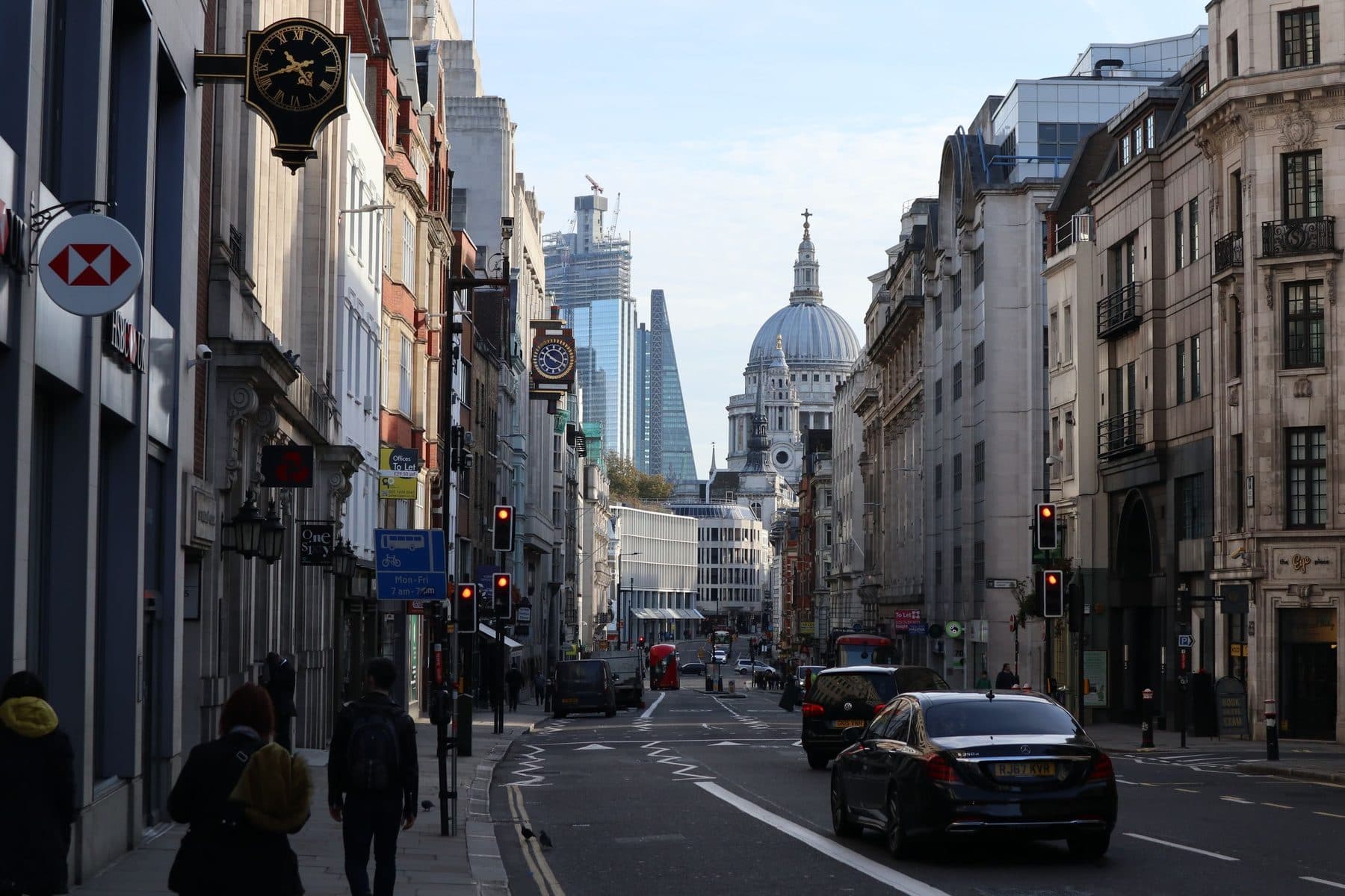 A London street of stone fronted buildings with a hanging clock, looking towards the dome of St Paul's Cathedral