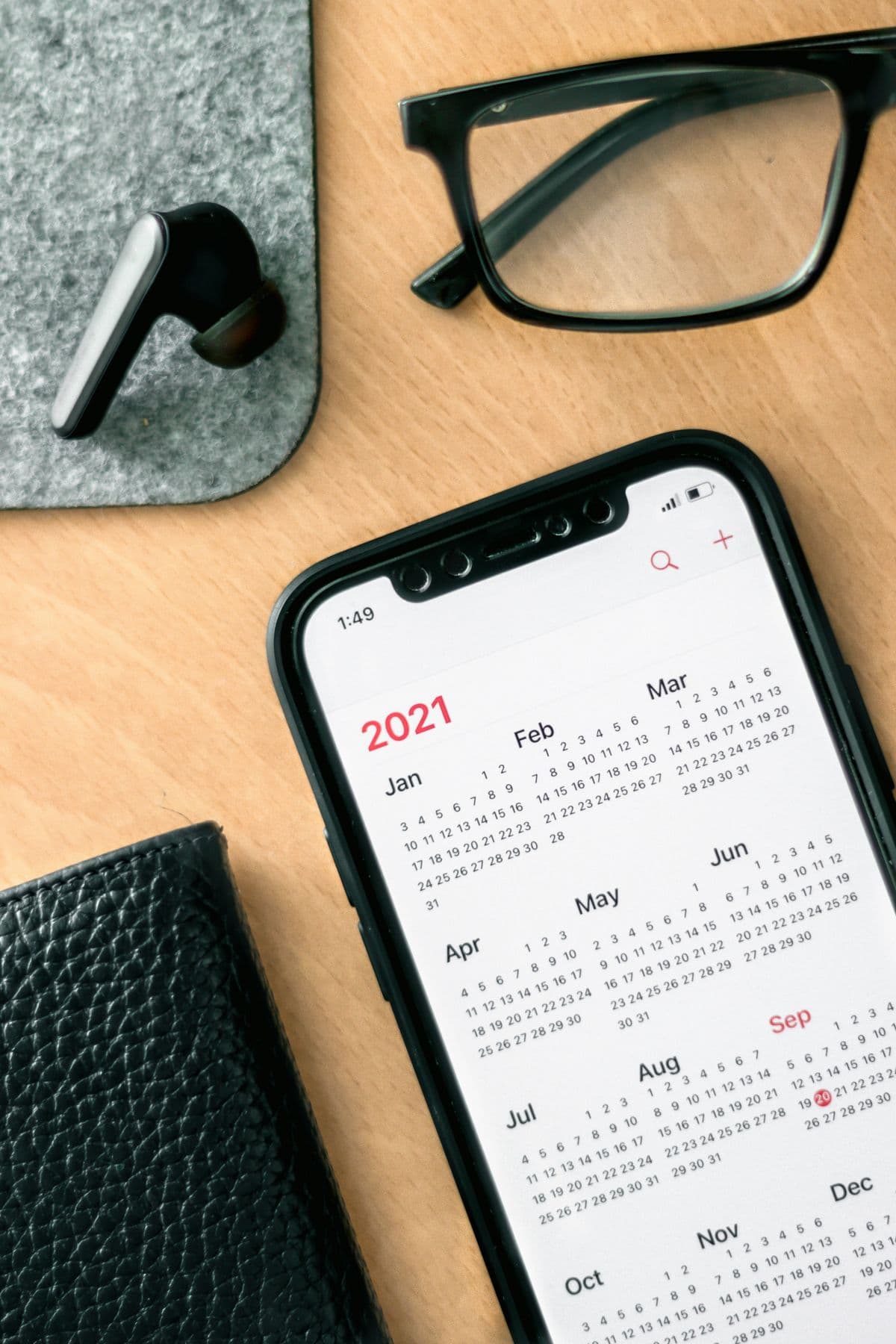 A phone showing a calendar beside a pair of glasses and earbuds on a warm wooden desk