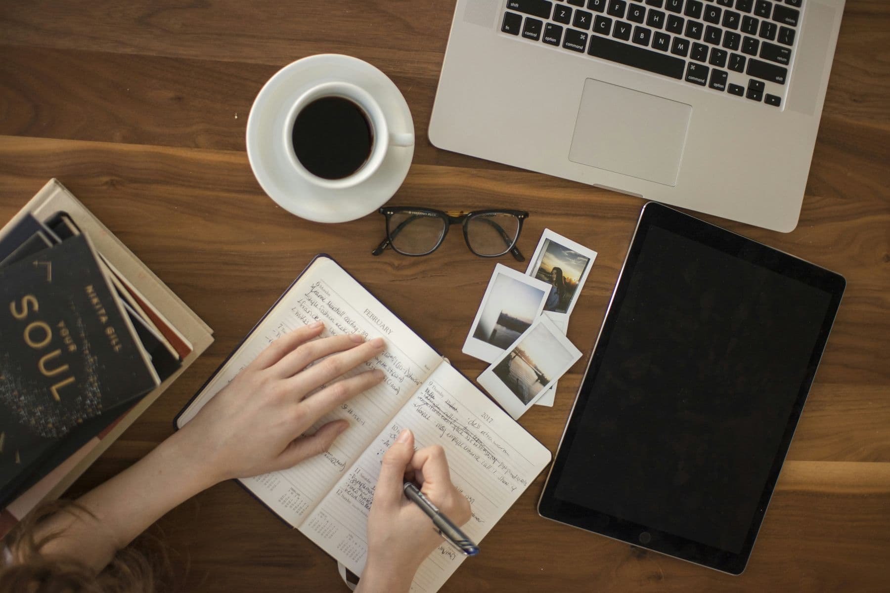 Hands writing notes in a notebook beside a laptop, a tablet, a coffee and a few printed photographs