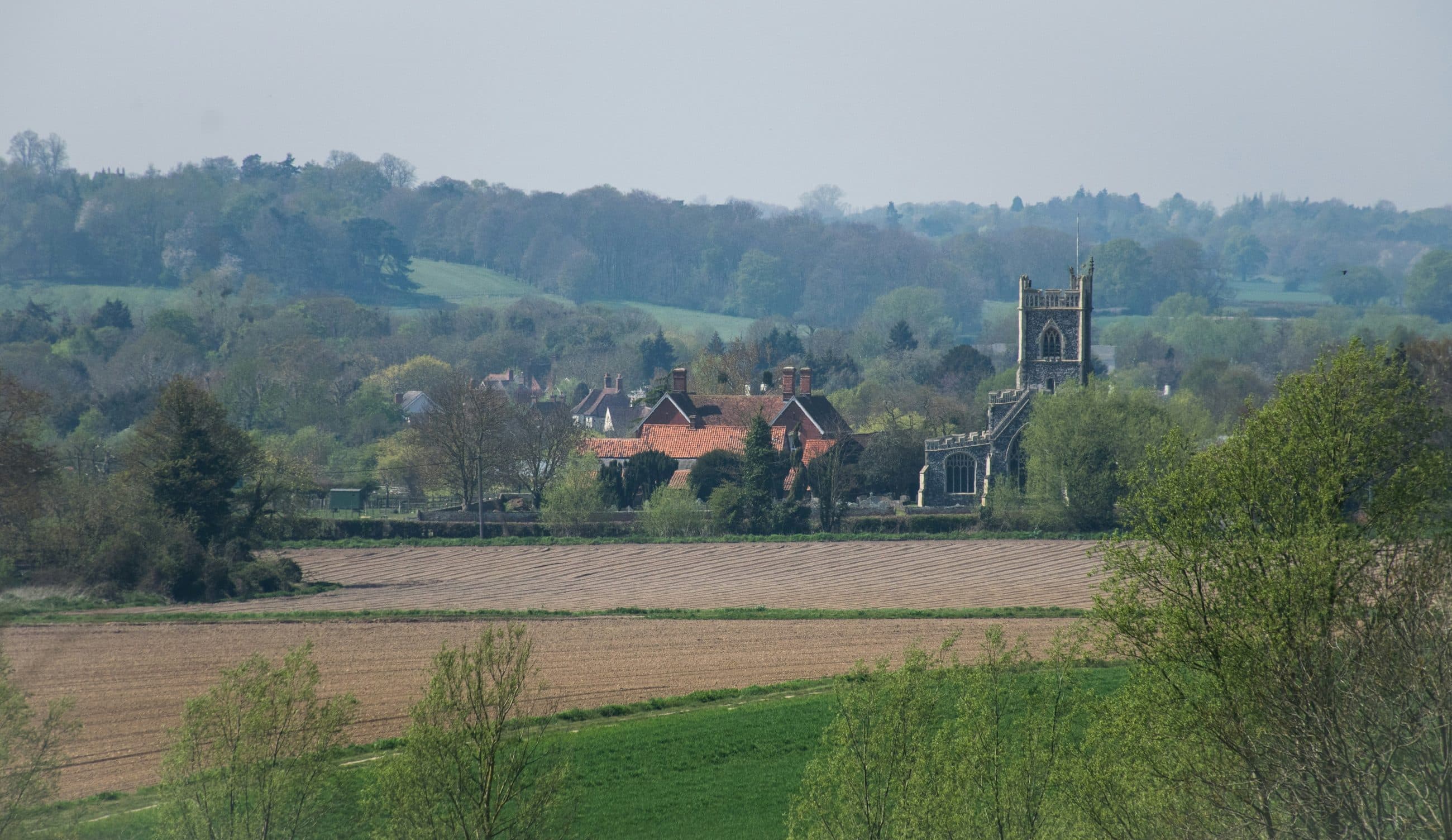 Dedham church tower and village roofs seen across ploughed fields and spring trees in the Dedham Vale, Essex