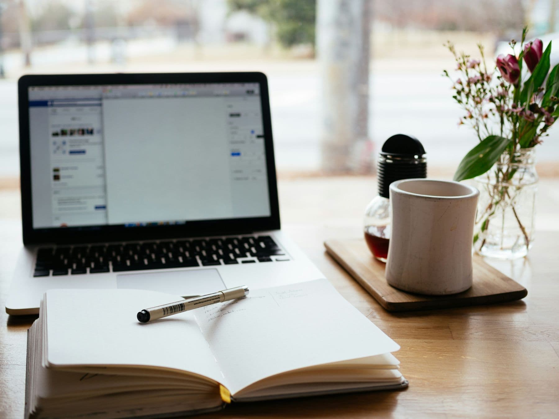 An open notebook and pen in front of a laptop, with a mug and a jar of flowers on a table by a window